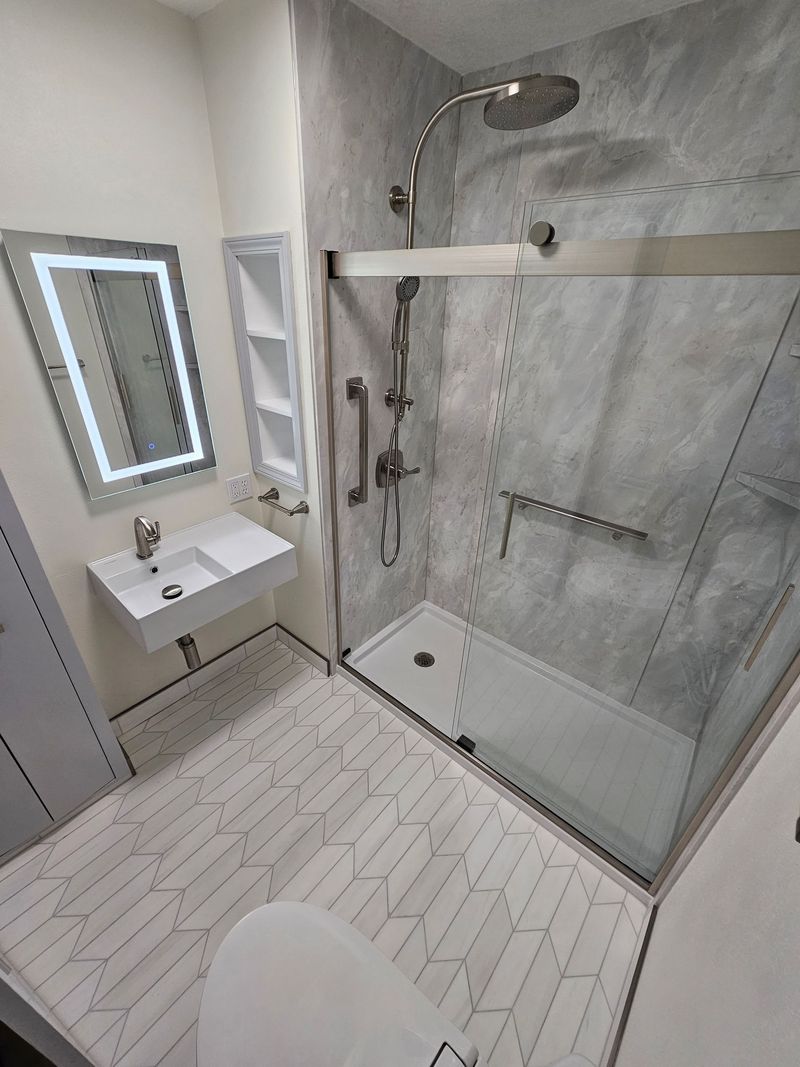 Walk-in shower with gray marble-look walls and glass doors beside a lighted mirror